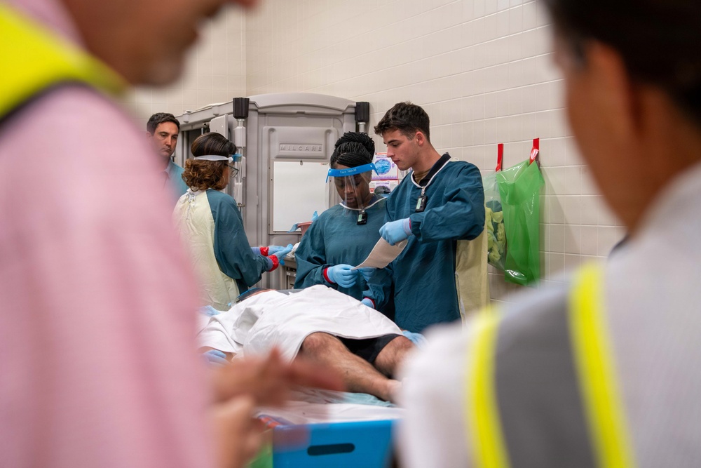 A medical team is attending to a patient lying on a gurney or examination table in what appears to be a clinical or emergency setting. Several healthcare workers wearing teal/blue scrubs, gloves, and at least one face shield are actively engaged with the patient and reviewing documents. Observers in high-visibility yellow vests are present in the foreground, suggesting this may be a training exercise or supervised medical scenario.