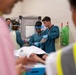 A medical team is attending to a patient lying on a gurney or examination table in what appears to be a clinical or emergency setting. Several healthcare workers wearing teal/blue scrubs, gloves, and at least one face shield are actively engaged with the patient and reviewing documents. Observers in high-visibility yellow vests are present in the foreground, suggesting this may be a training exercise or supervised medical scenario.