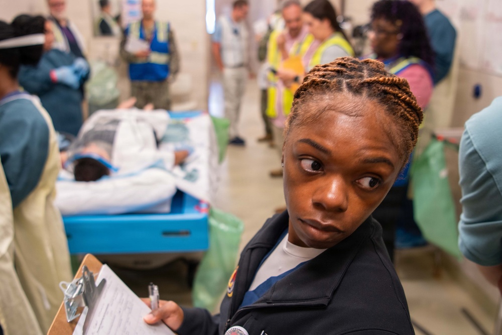 A woman in a dark uniform holding a clipboard with paperwork looks back toward the camera in what appears to be a medical triage or emergency response setting. Multiple personnel wearing high-visibility vests, blue vests, and medical gowns are visible in the background. A person appears to be lying on a gurney or cot in the mid-ground, suggesting an active medical or disaster response exercise.