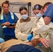 A medical team of five individuals is attending to a patient lying on a table, performing what appears to be a wound treatment or emergency medical procedure. Three clinicians in gowns, masks, and gloves are actively working on the patient's arm/torso area, while a person in scrubs observes and another in a high-visibility vest takes notes. The setting appears to be a medical training facility or emergency treatment room.