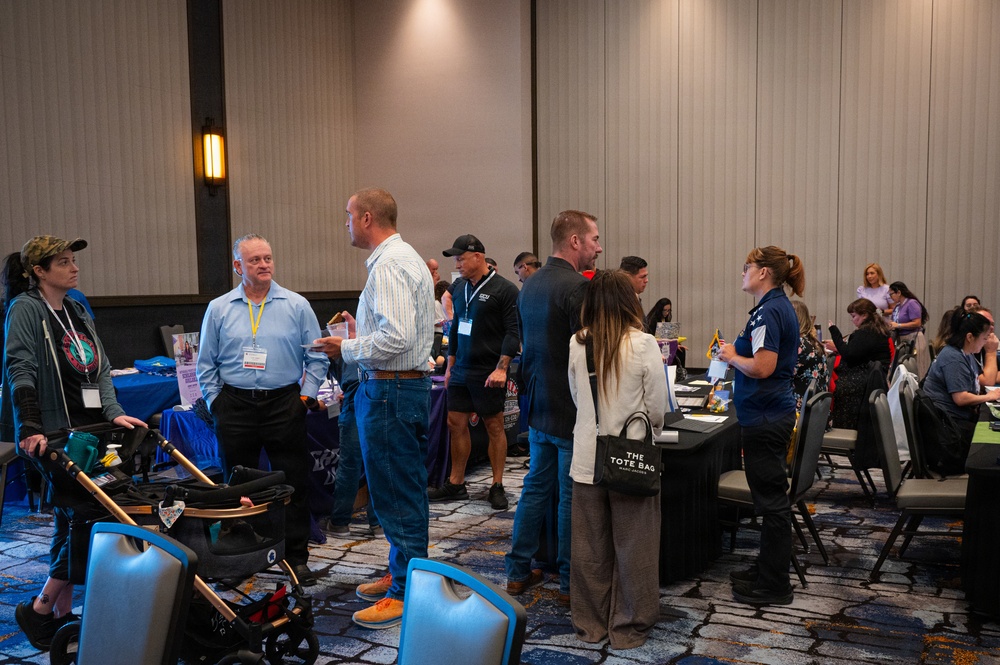 The image shows a busy indoor conference or expo event taking place in what appears to be a hotel ballroom or convention center. Attendees are mingling, browsing vendor or information tables, and engaging in conversations. The event appears to be a community or professional fair with multiple exhibitor booths.