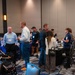 The image shows a busy indoor conference or expo event taking place in what appears to be a hotel ballroom or convention center. Attendees are mingling, browsing vendor or information tables, and engaging in conversations. The event appears to be a community or professional fair with multiple exhibitor booths.