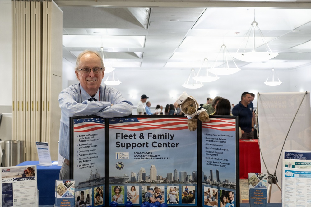 An older man with glasses smiles while leaning on a display booth for the Fleet &amp; Family Support Center, likely at a military resource fair or expo. The booth features informational signage, brochures, and a small stuffed animal mascot wearing a sailor hat. The background shows a large indoor venue with other attendees and booths visible.