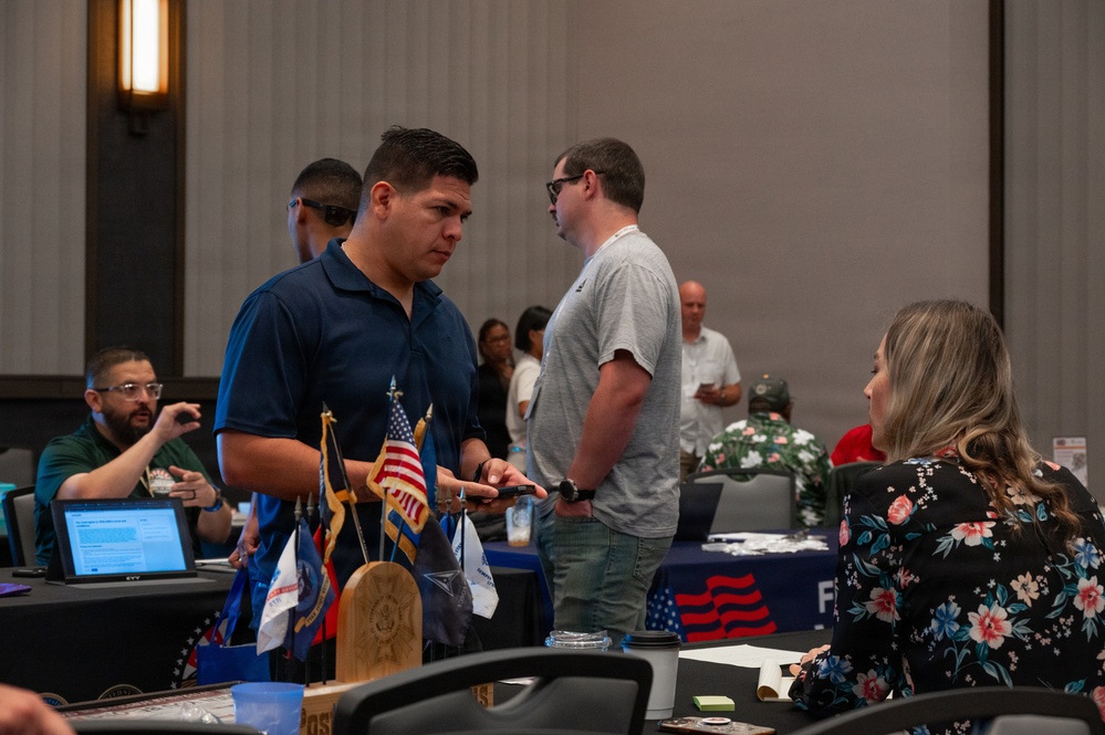 The image depicts what appears to be a veterans' resource fair or job fair held in a conference or banquet hall. Multiple attendees are engaged in conversations at various booths or tables, with American flags and patriotic decorations visible. The event appears to be organized to connect veterans or military-affiliated individuals with services or employment opportunities.