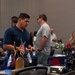 The image depicts what appears to be a veterans' resource fair or job fair held in a conference or banquet hall. Multiple attendees are engaged in conversations at various booths or tables, with American flags and patriotic decorations visible. The event appears to be organized to connect veterans or military-affiliated individuals with services or employment opportunities.