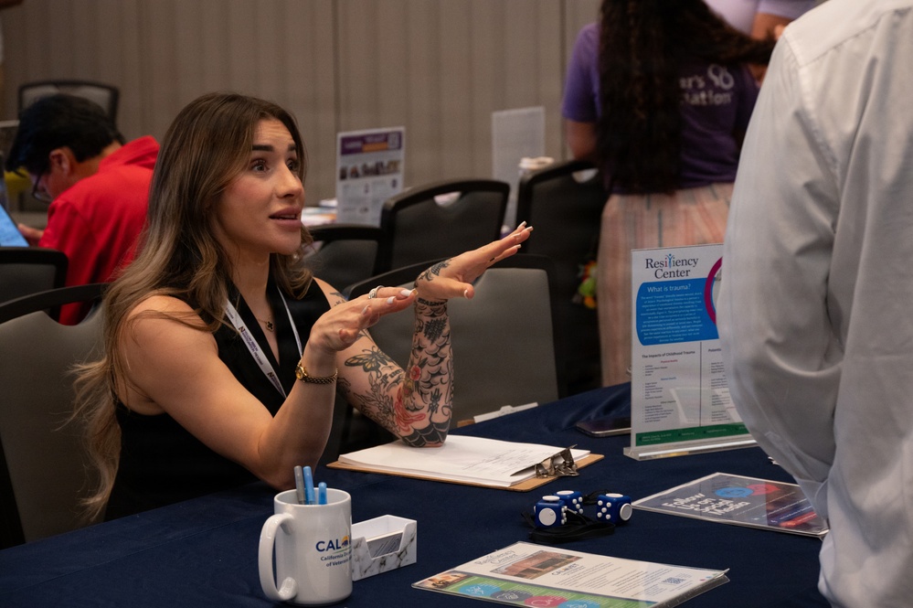A woman with visible tattoos on her arms sits at a booth for the 'Resiliency Center' at what appears to be a health or community services fair. She is actively speaking and gesturing with her hands toward a person standing across the table, likely explaining services. The table is covered with informational materials, a clipboard, dice, and a mug labeled 'CAL' (likely California).