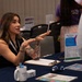 A woman with visible tattoos on her arms sits at a booth for the 'Resiliency Center' at what appears to be a health or community services fair. She is actively speaking and gesturing with her hands toward a person standing across the table, likely explaining services. The table is covered with informational materials, a clipboard, dice, and a mug labeled 'CAL' (likely California).