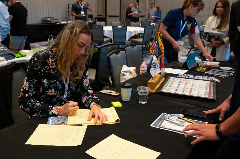 A woman in a floral dress is writing notes at a table during what appears to be a military or veterans-related conference or expo. The table is covered with informational materials, flags, and organizational literature. Multiple attendees and staff are visible in the background of what appears to be a hotel ballroom or convention center.