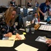 A woman in a floral dress is writing notes at a table during what appears to be a military or veterans-related conference or expo. The table is covered with informational materials, flags, and organizational literature. Multiple attendees and staff are visible in the background of what appears to be a hotel ballroom or convention center.