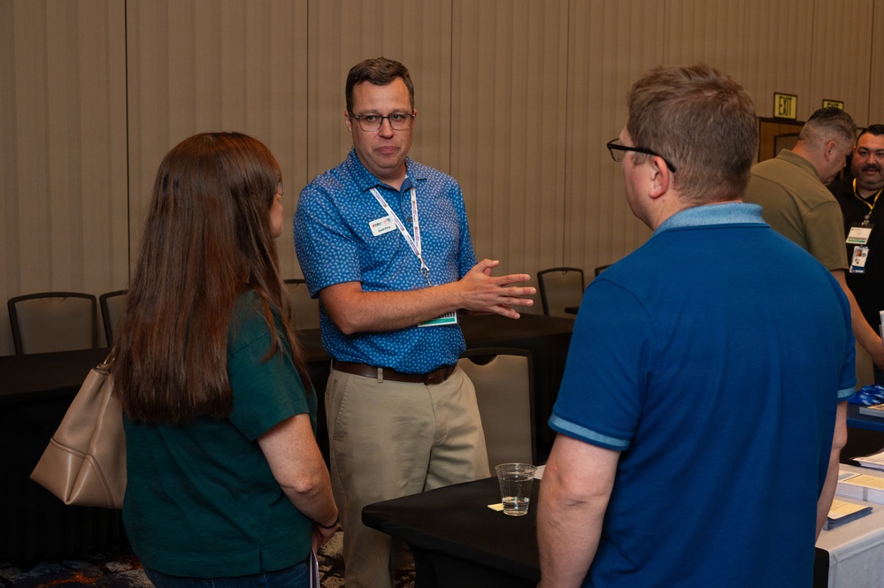 A professional networking or conference event taking place in what appears to be a hotel ballroom or convention center. A man wearing a blue patterned shirt, lanyard with name badge, and khaki pants is engaged in conversation with two other attendees. He is gesturing with his hand as if explaining or presenting something.