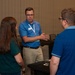 A professional networking or conference event taking place in what appears to be a hotel ballroom or convention center. A man wearing a blue patterned shirt, lanyard with name badge, and khaki pants is engaged in conversation with two other attendees. He is gesturing with his hand as if explaining or presenting something.