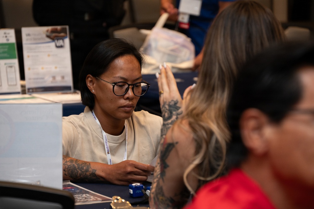 The image shows what appears to be a professional event or resource fair, likely related to the VA (Department of Veterans Affairs) based on visible signage. A person wearing glasses, a lanyard, and a cream-colored shirt with visible arm tattoos is seated at a table, appearing to assist or consult with another individual. The setting suggests an informational booth or service counter environment.