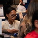 The image shows what appears to be a professional event or resource fair, likely related to the VA (Department of Veterans Affairs) based on visible signage. A person wearing glasses, a lanyard, and a cream-colored shirt with visible arm tattoos is seated at a table, appearing to assist or consult with another individual. The setting suggests an informational booth or service counter environment.