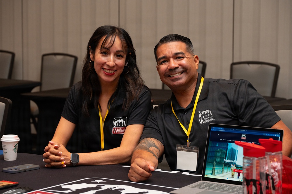 Two individuals wearing matching black 'Work for Warriors' polo shirts and yellow lanyards are seated at a table, smiling at the camera. They appear to be staffing an informational booth or recruitment table at a conference or event. A laptop displaying the Work for Warriors website, a Starbucks cup, red water bottles, and printed materials are visible on the table.