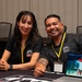 Two individuals wearing matching black 'Work for Warriors' polo shirts and yellow lanyards are seated at a table, smiling at the camera. They appear to be staffing an informational booth or recruitment table at a conference or event. A laptop displaying the Work for Warriors website, a Starbucks cup, red water bottles, and printed materials are visible on the table.