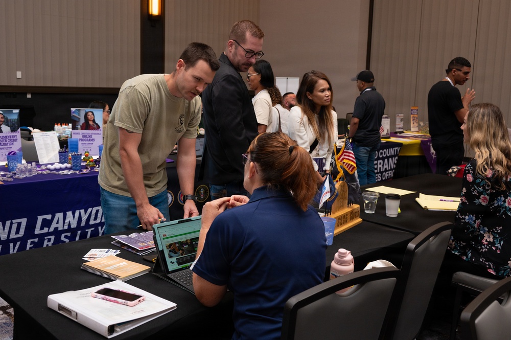 The image shows an indoor expo or resource fair, likely a veterans or military transition event, taking place in a conference or ballroom setting. Multiple booths are visible, including one for Grand Canyon University and what appears to be a County Veterans Services Office. Attendees are interacting with representatives at tables equipped with laptops, brochures, and informational materials.