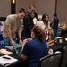 The image shows an indoor expo or resource fair, likely a veterans or military transition event, taking place in a conference or ballroom setting. Multiple booths are visible, including one for Grand Canyon University and what appears to be a County Veterans Services Office. Attendees are interacting with representatives at tables equipped with laptops, brochures, and informational materials.