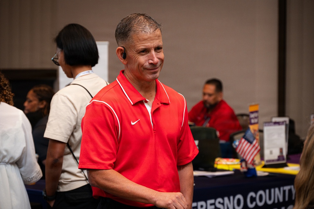 A middle-aged man wearing a red Nike polo shirt stands in the foreground at what appears to be a community or government event. He has short gray hair and is wearing a small earpiece. In the background, a 'Fresno County' branded table is visible with another person seated behind it, along with an American flag and various materials.