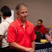 A middle-aged man wearing a red Nike polo shirt stands in the foreground at what appears to be a community or government event. He has short gray hair and is wearing a small earpiece. In the background, a 'Fresno County' branded table is visible with another person seated behind it, along with an American flag and various materials.