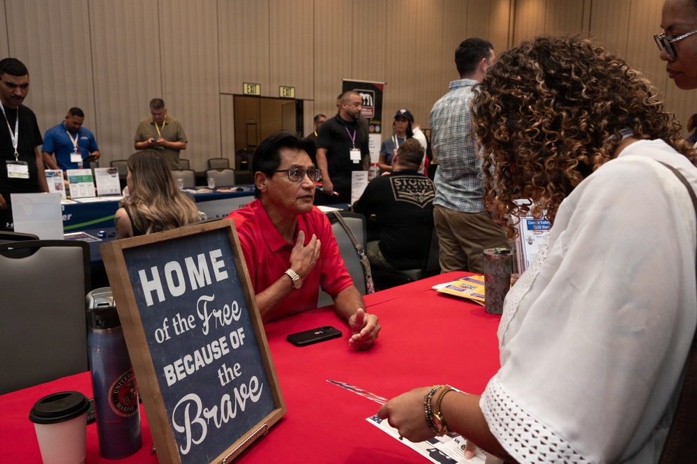 A man in a red shirt sits behind a red-draped table at what appears to be a veterans or military-themed job fair or expo, speaking with two women standing across from him. A chalkboard sign reading 'Home of the Free Because of the Brave' is prominently displayed on the table. The background shows a busy convention hall with multiple booths, attendees wearing lanyards, and various display materials.