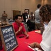 A man in a red shirt sits behind a red-draped table at what appears to be a veterans or military-themed job fair or expo, speaking with two women standing across from him. A chalkboard sign reading 'Home of the Free Because of the Brave' is prominently displayed on the table. The background shows a busy convention hall with multiple booths, attendees wearing lanyards, and various display materials.