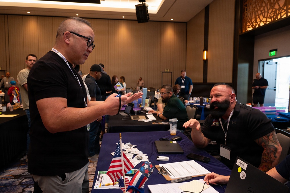 The image shows what appears to be a veterans-oriented job fair or resource event held in a hotel ballroom or conference center. A man wearing a lanyard badge is speaking with a seated representative at a table draped in a blue tablecloth, which displays American flags and various documents. Multiple similar booths and attendees are visible in the background.