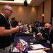 The image shows what appears to be a veterans-oriented job fair or resource event held in a hotel ballroom or conference center. A man wearing a lanyard badge is speaking with a seated representative at a table draped in a blue tablecloth, which displays American flags and various documents. Multiple similar booths and attendees are visible in the background.
