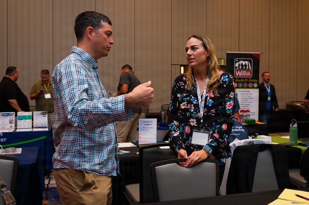 Two individuals are engaged in conversation at what appears to be a military or veterans-focused career/resource fair. The woman is wearing a conference lanyard and floral dress, while the man in a plaid shirt gestures as he speaks. Multiple vendor booths are visible in the background, including 'Work for Warriors' and 'Soldiers' Angels'.
