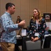 Two individuals are engaged in conversation at what appears to be a military or veterans-focused career/resource fair. The woman is wearing a conference lanyard and floral dress, while the man in a plaid shirt gestures as he speaks. Multiple vendor booths are visible in the background, including 'Work for Warriors' and 'Soldiers' Angels'.