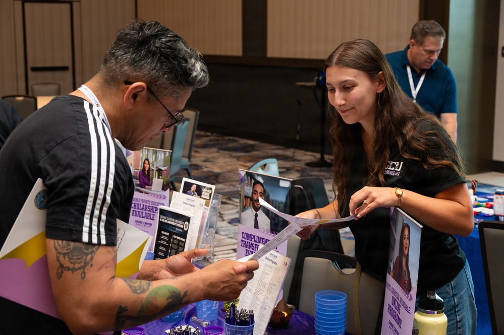 A young woman wearing a black GCU (Grand Canyon University) shirt is presenting informational materials to a man with tattoos and glasses at what appears to be an educational fair or recruitment event. The table displays multiple brochures and promotional materials for Grand Canyon University, including signs about scholarships, online programs, and transfer evaluations. A third person wearing a lanyard is visible in the background.