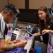 A young woman wearing a black GCU (Grand Canyon University) shirt is presenting informational materials to a man with tattoos and glasses at what appears to be an educational fair or recruitment event. The table displays multiple brochures and promotional materials for Grand Canyon University, including signs about scholarships, online programs, and transfer evaluations. A third person wearing a lanyard is visible in the background.