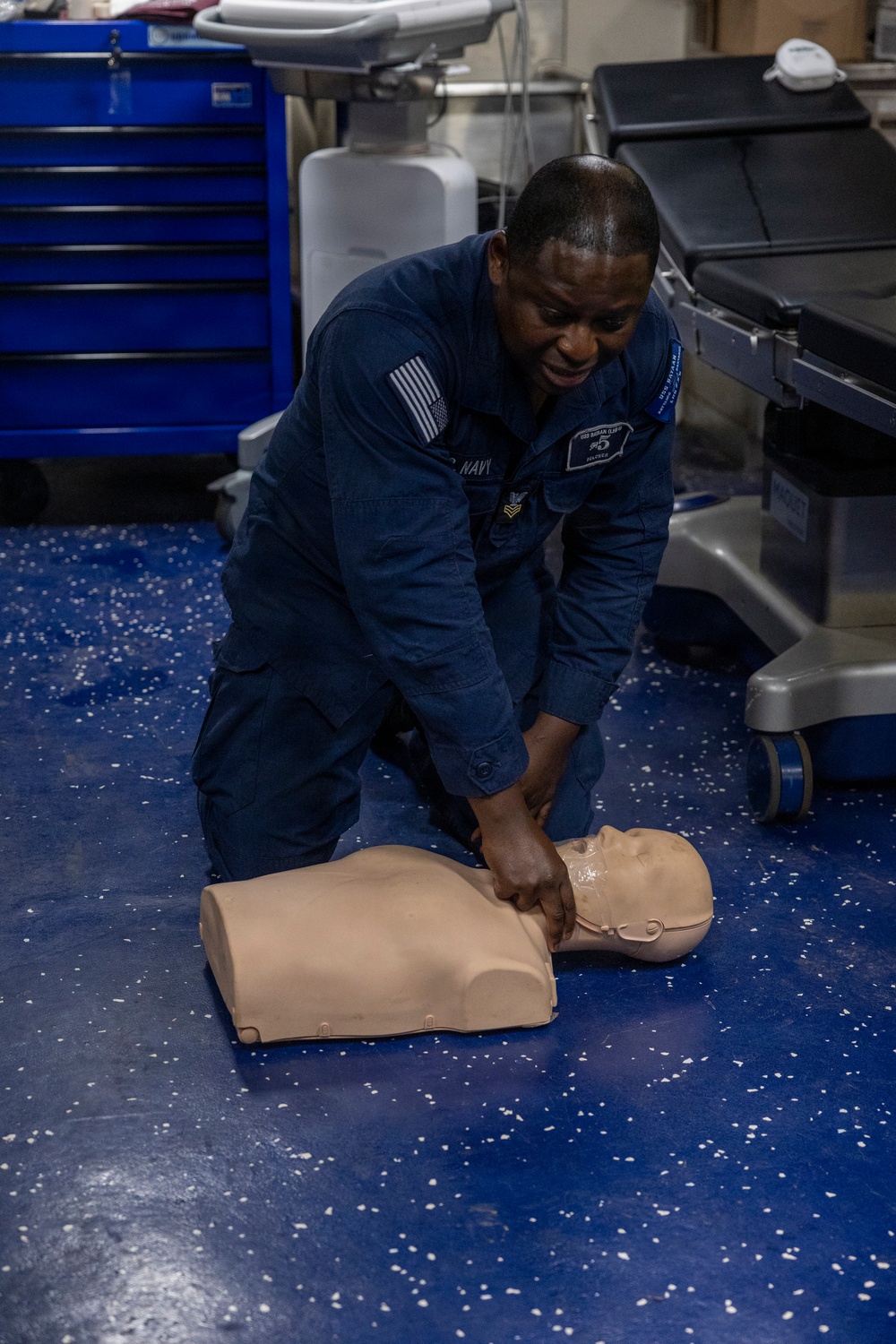 A U.S. Navy sailor in blue utility uniform is kneeling on a blue speckled floor performing chest compressions on a CPR training manikin. The setting appears to be a medical or sick bay facility aboard a naval vessel, indicated by the medical equipment visible in the background. This appears to be a CPR/first aid training exercise.