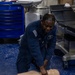 A U.S. Navy sailor in blue utility uniform is kneeling on a blue speckled floor performing chest compressions on a CPR training manikin. The setting appears to be a medical or sick bay facility aboard a naval vessel, indicated by the medical equipment visible in the background. This appears to be a CPR/first aid training exercise.