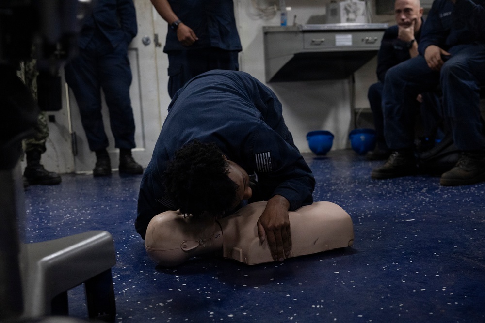 A person in dark blue military/naval uniform is performing CPR rescue breathing on a training mannequin on a blue floor. This appears to be a CPR/first aid training session, with several other uniformed personnel observing in the background. Blue safety helmets are visible on the floor nearby, suggesting a military or maritime training environment.