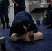A person in dark blue military/naval uniform is performing CPR rescue breathing on a training mannequin on a blue floor. This appears to be a CPR/first aid training session, with several other uniformed personnel observing in the background. Blue safety helmets are visible on the floor nearby, suggesting a military or maritime training environment.