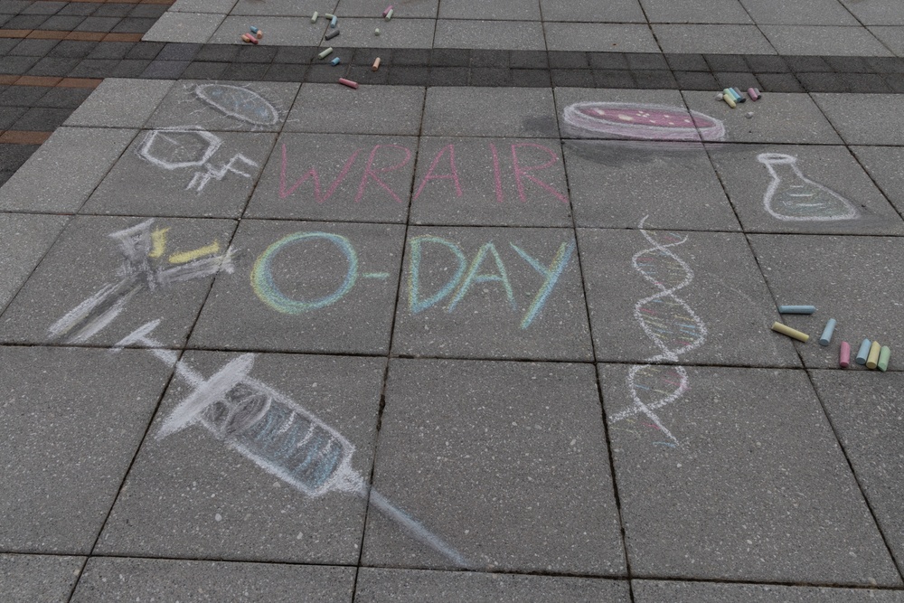 Chalk art drawn on a paved sidewalk or plaza promoting 'WRAIR O-Day,' which refers to the Walter Reed Army Institute of Research Orientation Day. The artwork features science and medical-themed illustrations drawn in multicolored chalk. Loose chalk pieces are scattered around the drawings on the pavement.