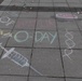 Chalk art drawn on a paved sidewalk or plaza promoting 'WRAIR O-Day,' which refers to the Walter Reed Army Institute of Research Orientation Day. The artwork features science and medical-themed illustrations drawn in multicolored chalk. Loose chalk pieces are scattered around the drawings on the pavement.