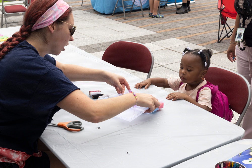 An adult woman with red braided hair and sunglasses is conducting a craft activity with a young toddler girl at an outdoor event. The child, wearing a pink backpack, is seated at a white folding table and appears engaged as the woman works with paper and craft materials. The setting appears to be a community or school fair held on a paved plaza.
