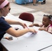 An adult woman with red braided hair and sunglasses is conducting a craft activity with a young toddler girl at an outdoor event. The child, wearing a pink backpack, is seated at a white folding table and appears engaged as the woman works with paper and craft materials. The setting appears to be a community or school fair held on a paved plaza.