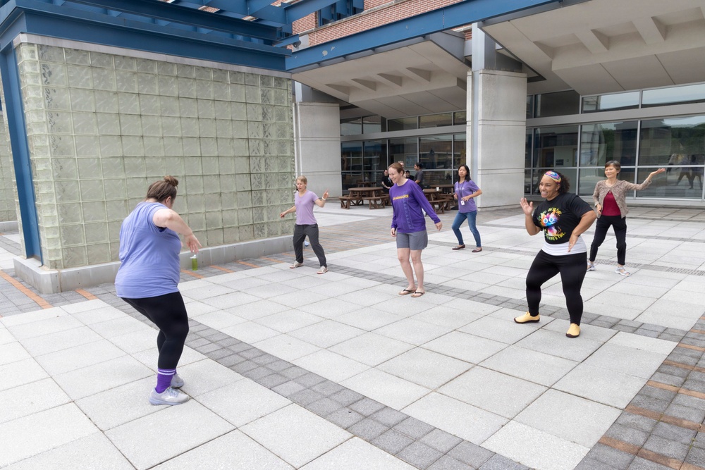A group of approximately six women are participating in an outdoor dance or exercise class on a paved plaza area adjacent to a modern building. The participants are spread out in an open courtyard, following along with movements that appear to be a fitness or Zumba-style activity. The setting appears to be a public or institutional facility, possibly a community center or university campus.