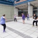 A group of approximately six women are participating in an outdoor dance or exercise class on a paved plaza area adjacent to a modern building. The participants are spread out in an open courtyard, following along with movements that appear to be a fitness or Zumba-style activity. The setting appears to be a public or institutional facility, possibly a community center or university campus.
