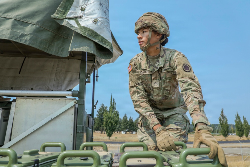 A U.S. Army soldier, identified by name tape as 'DAZLE', is working with military equipment in an outdoor setting. The soldier is wearing full OCP (Operational Camouflage Pattern) uniform with helmet, tactical gloves, and has face camouflage paint applied. The setting appears to be a military training or operational area with green military vehicles and equipment visible.