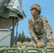A U.S. Army soldier, identified by name tape as 'DAZLE', is working with military equipment in an outdoor setting. The soldier is wearing full OCP (Operational Camouflage Pattern) uniform with helmet, tactical gloves, and has face camouflage paint applied. The setting appears to be a military training or operational area with green military vehicles and equipment visible.