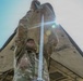 A soldier in MultiCam/OCP camouflage uniform is photographed from a low-angle perspective while erecting or adjusting a military tent. The dramatic upward camera angle, combined with direct sun flare, creates a striking compositional effect showing the tent canopy framing the soldier against the sky. The soldier is using both gloved hands to manipulate a metal tent pole at the apex of the structure.