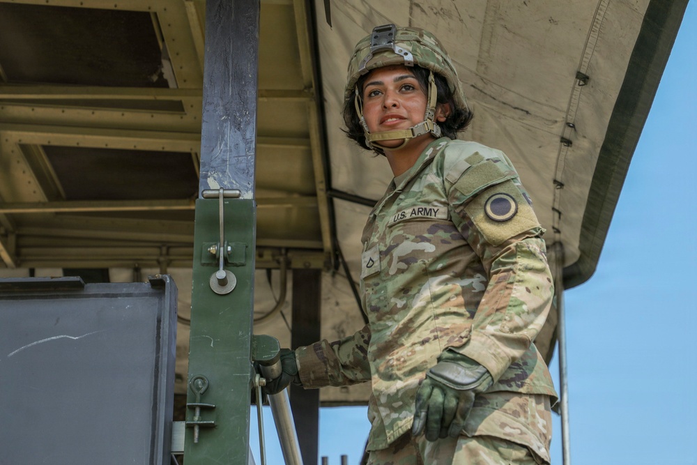 A U.S. Army soldier in full Operational Camouflage Pattern (OCP) uniform and Advanced Combat Helmet is photographed from a low angle while standing on or near a military vehicle. The soldier is wearing tactical gloves and gripping a metal handle or lever on what appears to be a large military transport or equipment platform. The image has a professional, portrait-style composition with a clear blue sky in the background.
