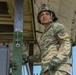 A U.S. Army soldier in full Operational Camouflage Pattern (OCP) uniform and Advanced Combat Helmet is photographed from a low angle while standing on or near a military vehicle. The soldier is wearing tactical gloves and gripping a metal handle or lever on what appears to be a large military transport or equipment platform. The image has a professional, portrait-style composition with a clear blue sky in the background.