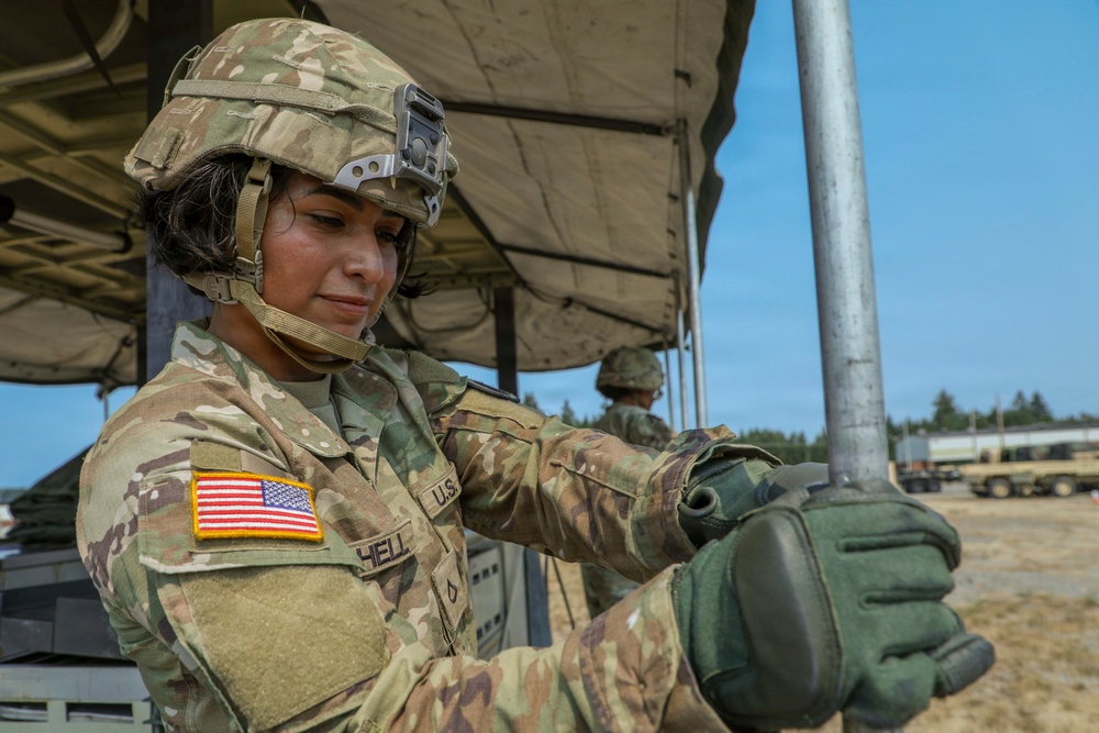 A U.S. Army soldier in full combat uniform and helmet is working with a metal pole or antenna component under a field canopy structure. The soldier, identified by a partial name tape reading 'HELL' and wearing an American flag patch on the left shoulder, appears to be assembling or adjusting equipment at what looks like a field operations site. A second soldier is visible in the background near military vehicles.