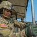 A U.S. Army soldier in full combat uniform and helmet is working with a metal pole or antenna component under a field canopy structure. The soldier, identified by a partial name tape reading 'HELL' and wearing an American flag patch on the left shoulder, appears to be assembling or adjusting equipment at what looks like a field operations site. A second soldier is visible in the background near military vehicles.