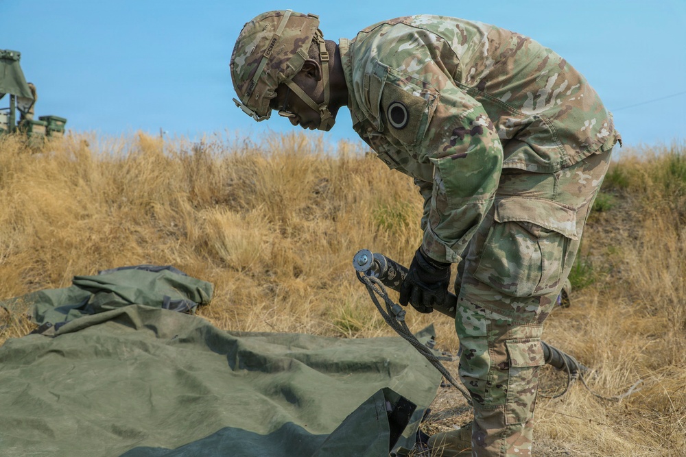 A U.S. Army soldier in Operational Camouflage Pattern (OCP) uniform is bent over handling rigging equipment, specifically a cable or strap with a metal hook/connector, near a green canvas tarp or equipment cover on the ground. The setting appears to be an outdoor field training environment with dry grassland in the background. Military equipment is partially visible in the upper left background.