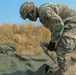 A U.S. Army soldier in Operational Camouflage Pattern (OCP) uniform is bent over handling rigging equipment, specifically a cable or strap with a metal hook/connector, near a green canvas tarp or equipment cover on the ground. The setting appears to be an outdoor field training environment with dry grassland in the background. Military equipment is partially visible in the upper left background.